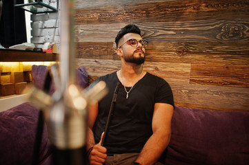 Stylish beard arabian man in glasses and black t-shirt smoking hookah indoor bar. Arab model having...