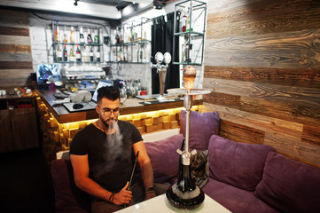 Stylish beard arabian man in glasses and black t-shirt smoking hookah indoor bar. Arab model having rest.