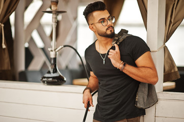 Stylish beard arabian man in glasses and black t-shirt smoking hookah outdoor. Arab model having...