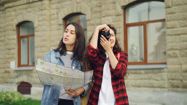 Female tourist is taking photos while her friend attractive girl is looking at map and showing her direction standing in the street then walking away.