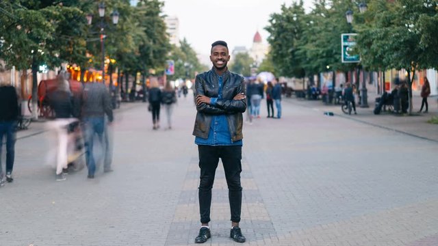 Time-lapse portrait of cheerful African American man standing in city center wearing stylish clothes looking at camera and smiling while people are passing by.