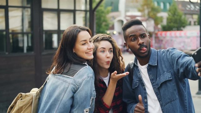 Good-looking young people Caucasian and African American tourists are taking selfie using smartphone posing with hand gestures having fun on summer day.