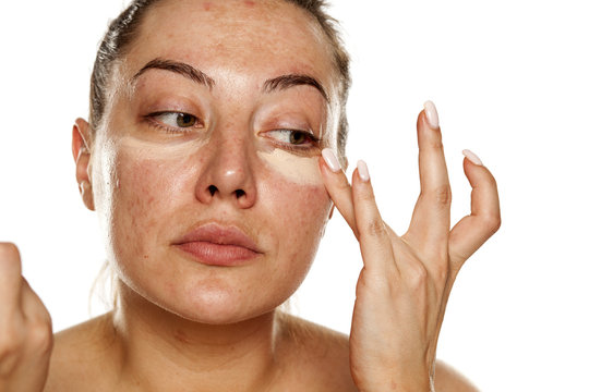Young woman applying concealer under her eyes with her fingers on white background