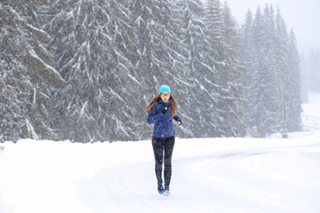 Fototapeta na wymiar Young woman running on the mountain road in bad weather in winter. Beautiful girl jogging in blizzard or snowstorm