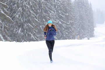 Fototapeta na wymiar Young woman running on the mountain road in bad weather in winter. Beautiful girl jogging in blizzard or snowstorm