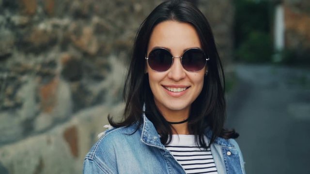 Close-up slow motion portrait of cute girl with dark hair in stylish sunglasses standing outside with stone wall in background, smiling and looking at camera.