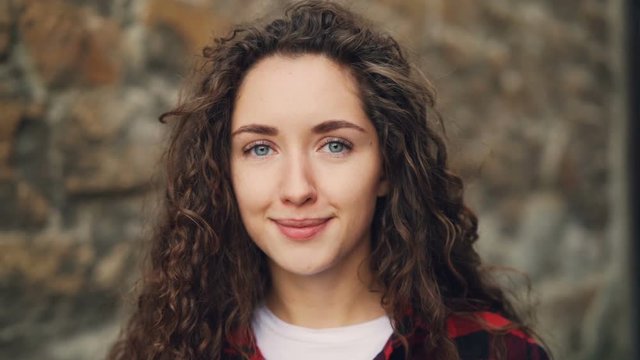Close-up slow motion portrait of pretty girl brunette with curly hair looking at camera with serious face then smiling reacting to good news. People and feelings concept.