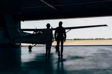 rear view of silhouettes of stylish couple walking near airplane in hangar - obrazy, fototapety, plakaty