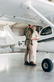 young fashionable couple in jacket embracing near plane in hangar