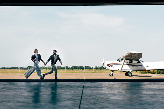 distant view of stylish young couple in leather jackets holding hands and running near plane - Powered by Adobe