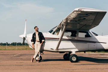 young fashionable male pilot in leather jacket and sunglasses posing near plane