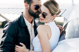 stylish man in leather jacket and sunglasses embracing attractive girlfriend near plane