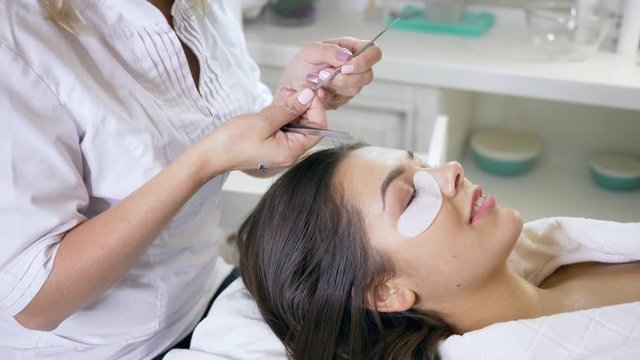 master and client in a beauty salon, professional stylist lengthening female lashes using special glue and two tweezers