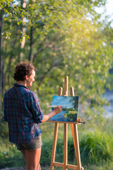 Fototapeta premium Young pretty woman artist draws paints a picture of a lake on open plain air outdoors