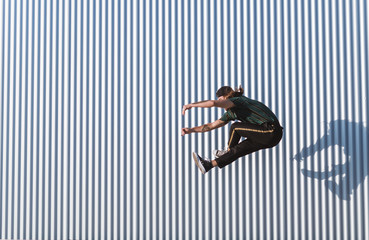 Young man with a cap and beard jumping in an acrobatics way on a sunny day of summer background an iron wall