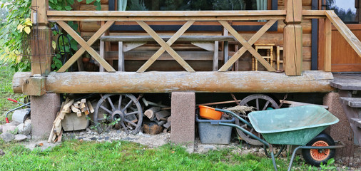 Under the porch of a wooden village shed is stored household utensils, firewood, basins and a wheelbarrow