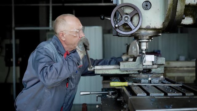 Old experienced plant worker in protective uniform drilling a piece of metal on special machine.