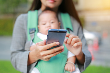 Close-up business mother in suit is using smartphone with carrying her baby boy in hipseat. Mother working with Baby.
