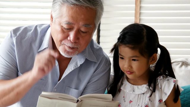 Senior man and little girl reading book together at home. people with family, lifestyle, education concept. 4k resolution.