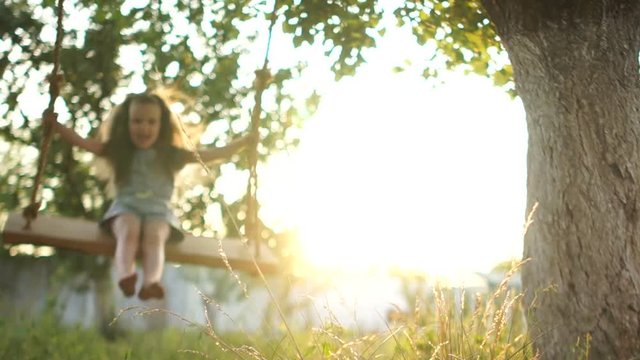 A barefoot girl rides on a wooden swing in the rays of the setting sun. Bottom view, close-up
