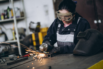Strong and worthy woman doing hard job in car and motorcycle repair shop. She using grinder to fix...