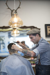 Barbero moderno con vestimenta vintage corta el pelo a joven muchacho veiteañero en barbería.