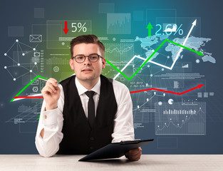 Young handsome businessman sitting at a desk with stocks and progress charts behind him