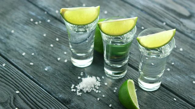 Row of glass shots with tequila and green sour lime slices on wooden table with heap of salt