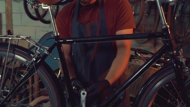 theme small business bike repair. young Caucasian brunette man wearing safety goggles, gloves and apron uses hand tool repairing and adjusting crank and pedal system bicycle in the garage workshop