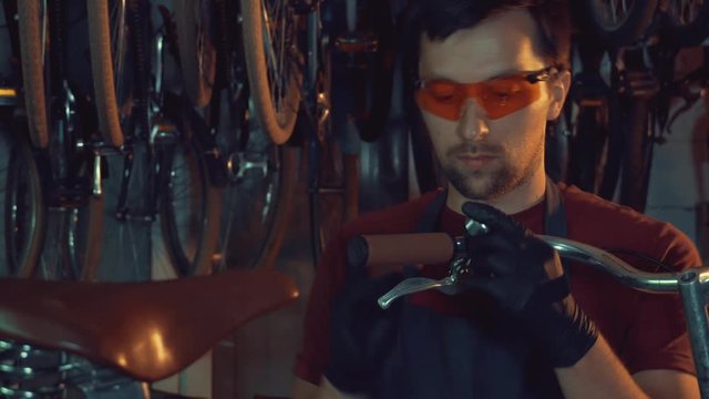 theme small business bike repair. A young Caucasian brunette man wearing safety goggles, gloves and apron uses hand tool to repair and adjust the Handbrabar brake bike handle in garage of workshop