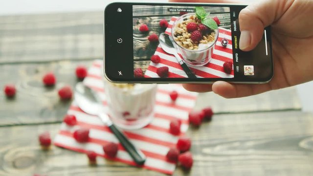Hand of anonymous person holding modern smartphone and making lovely photo of delicious fruit dessert and spoon on wooden table