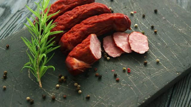 Twig of fresh rosemary and seeds of pepper lying on scratched board near delicious sliced and whole sausages