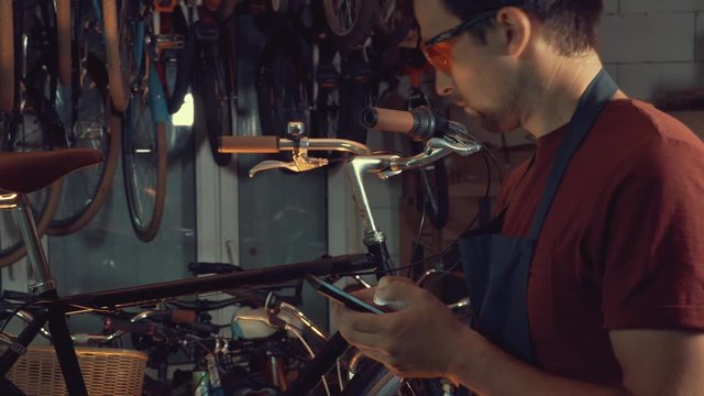 theme small business bike repair. Young caucasian brunette man wearing safety goggles, gloves and fartukhe uses mobile phone technology, takes notes, checklist in bicycle workshop