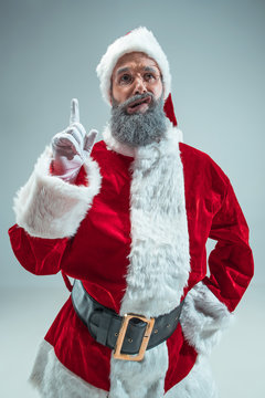 Funny guy with christmas hat posing at studio pointing up. New Year Holiday. Christmas, x-mas, winter, gifts concept. Man wearing Santa Claus costume on gray. Copy space. Winter sales.
