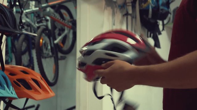 A young Caucasian man stands near a stand in a bicycle store in the head protection department. Choosing a bicycle helmet in a small store