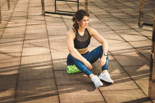 theme sport and rehabilitation sports medicine. Beautiful strong slender Caucasian woman athlete sits next foam roller green field street workout to remove the pain, stretch and massage muscles