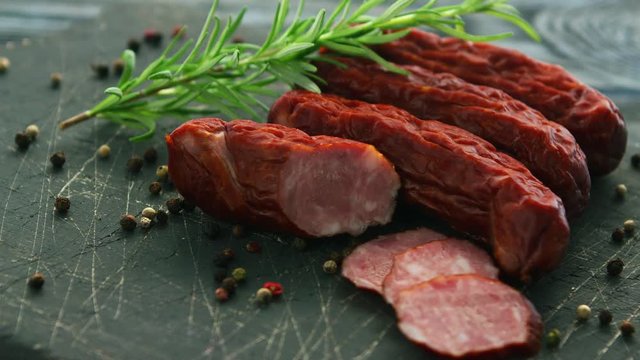 Closeup shot of delectable smoked sausages lying near twig of aromatic rosemary and seeds of hot pepper on scratched cutting board