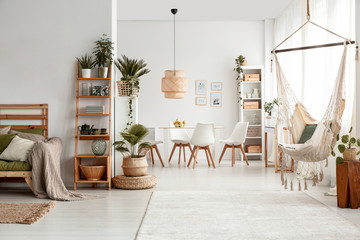 Hammock and plants in white apartment interior with lamp above dining table and chairs. Real photo