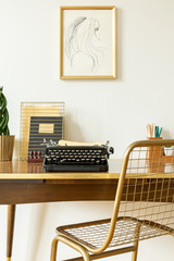 Industrial, golden net chair by a wooden desk with a black, vintage typewriter in an artistic home office interior