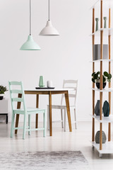 Lamps above wooden table and chairs in white and green dining room interior with shelves. Real photo