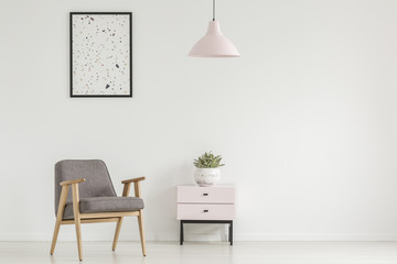 Poster above grey wooden armchair next to cabinet with plant in white flat interior with lamp. Real...