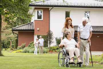 An elderly woman on a wheelchair and her husband with a cane with their medical assistant outside in the garden of a private care home.