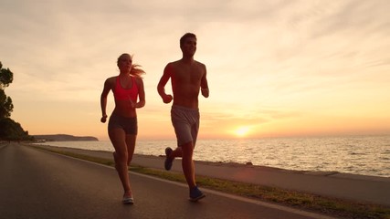SLOW MOTION, LENS FLARE: Happy fit couple jogging down the coastal road on a picturesque summer evening. Smiling girlfriend and boyfriend run shirtless along the beautiful coast at colorful sunset.