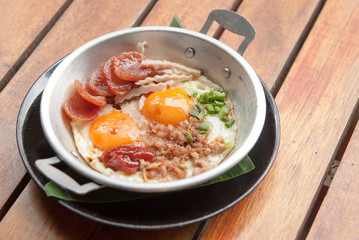 Morning breakfast, fried egg topped with minced pork, pork Thai sausage small sliced and vegetable in an aluminum pan on wooden table together warm light in the morning