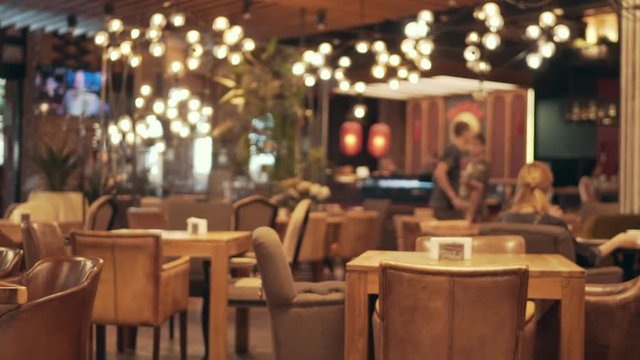 Blurred picture of the interior of a large beautiful restaurant with bright lighting, of the entrance. Tables with chairs are waiting for visitors for dinner. Visitors choose their own table

