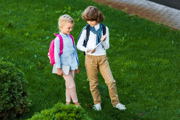 high angle view of children with backpacks holding digital tablet while standing together on green lawn