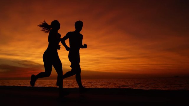 SILHOUETTE: Athletic young male and female workout partners jogging down the scenic road at romantic sunset. Healthy couple exercising during their summer vacation on a picturesque orange lit evening.