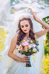 Beautiful bride in fashion wedding dress on natural background. The stunning young bride with long wedding veil is incredibly happy. Wedding day. A beautiful bride portrait in the green park. 