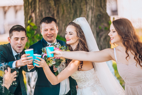 Newlyweds And Their Friends Open Champagne Bottle Standing In The Newlyweds And Their Friends Open Champagne Bottle Standing In The Park Before Wedding Ceremony