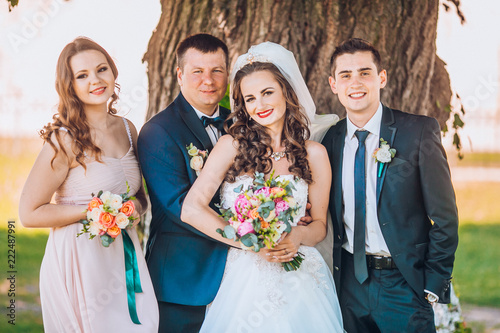 Newlyweds And Their Friends Open Champagne Bottle Standing In The Newlyweds And Their Friends Open Champagne Bottle Standing In The Park Before Wedding Ceremony